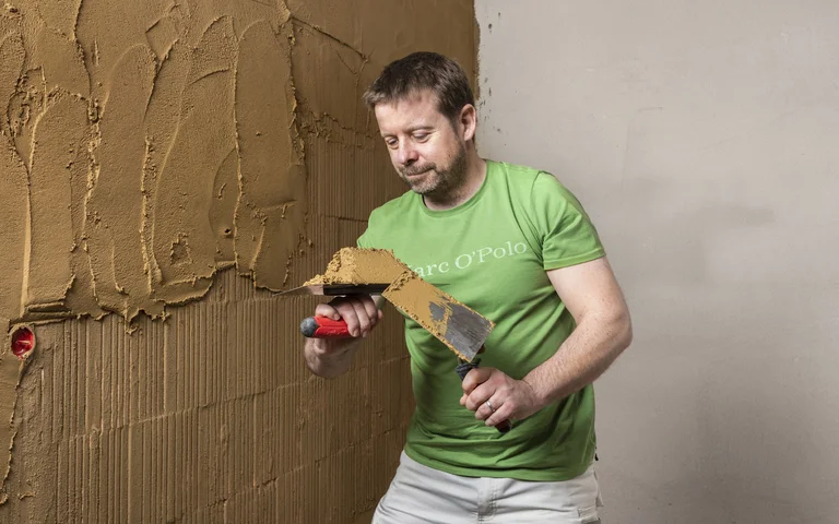 Person trägt mit Hilfe einer Kelle den Lehmputz an der Wand im Innenbereich auf.
