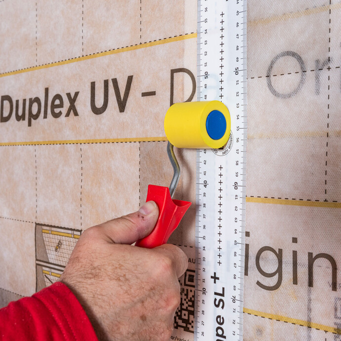 Das Vario MultiTape SL+ wird beidseitig gleichmäßig überlappend auf die Stoßbereiche zwischen Folienstreifen der Laibung und der bereits angebrachten Dampfbremse an der Wand geklebt. Mit einer Andruckwalze klebst du das Klebeband fest an.