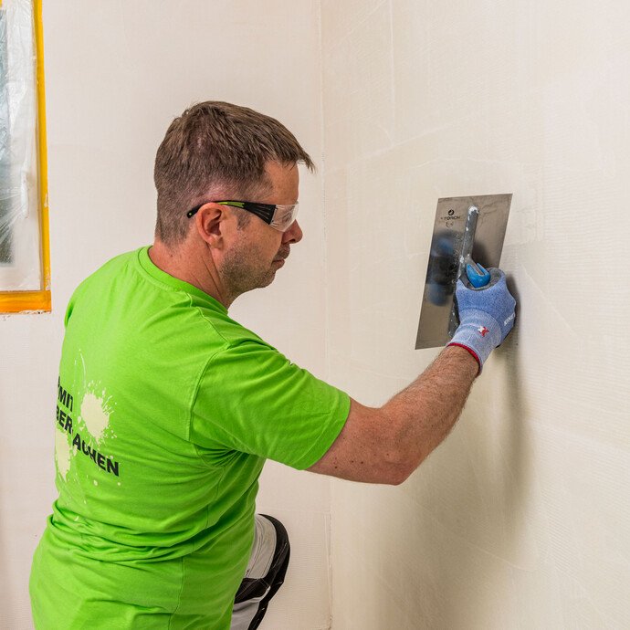 eimwerker kratzt abgebundenen Baumit Kalkin Kalkspachtel mit der scharfen Kante der Glättkelle auf – Grate und Unebenheiten entfernen für eine griffige Oberfläche vor der Deckspachtelung, Baumit Selbermachen Anleitung.
