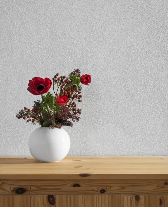 Holzregal mit weißer Vase und roten Blumen vor weißer, verputzter Wand.