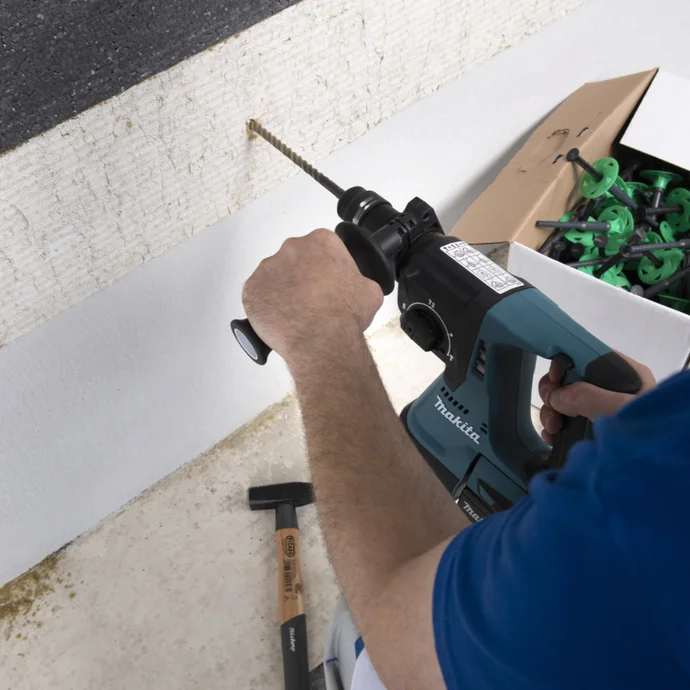 Person bohrt Loch in eine Wand zur Befestigung von Dämmstoff, Werkzeug und Dübel liegen bereit, Schritt zur Fassadensanierung.