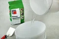 Wasser wird in einen Eimer gegossen, davor Baumit Innenspachtel, eine Kelle und Spachtelmasse für DIY-Projekte.