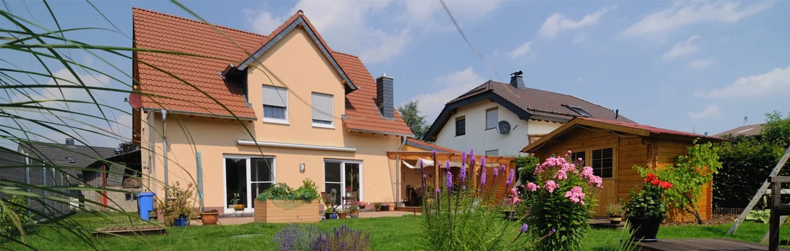 Haus mit gelber Fassade und Holzschuppen, umgeben von bl&uuml;henden G&auml;rten, ideal f&uuml;r Heimwerkerprojekte mit Baumit-Produkten.
