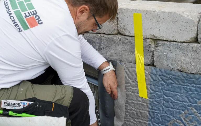Der Handwerker verklebt die beiden anstoßenden Noppenbahnen miteinander an der Gartenmauer. Rechts ist die Noppenbahn mit einem gelben Klebestreifen fixiert.