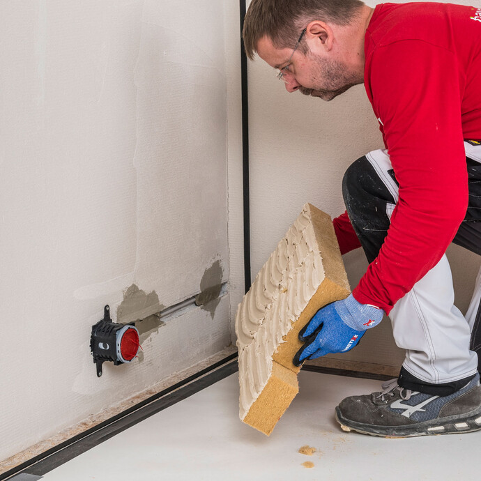 Heimwerker setzt mit Baumit Klebemörtel beschichtete Holzfaserdämmplatte passgenau an der Innendämmungsdose an – Dämmplatte mit Steckdosenaussparung sauber und vollflächig an die Wand kleben, Baumit Selbermachen Anleitung.