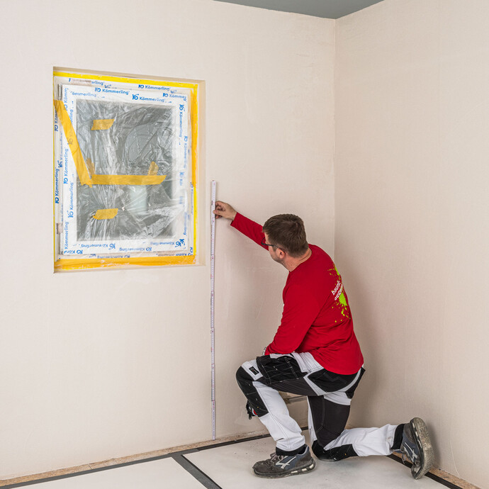 Heimwerker misst mit einem Zollstock die Höhe der Wandfläche neben einem abgedeckten Fenster aus – Wandhöhe prüfen vor dem Kleben der Baumit Holzfaserdämmplatten als Innendämmung, Baumit Selbermachen Anleitung.