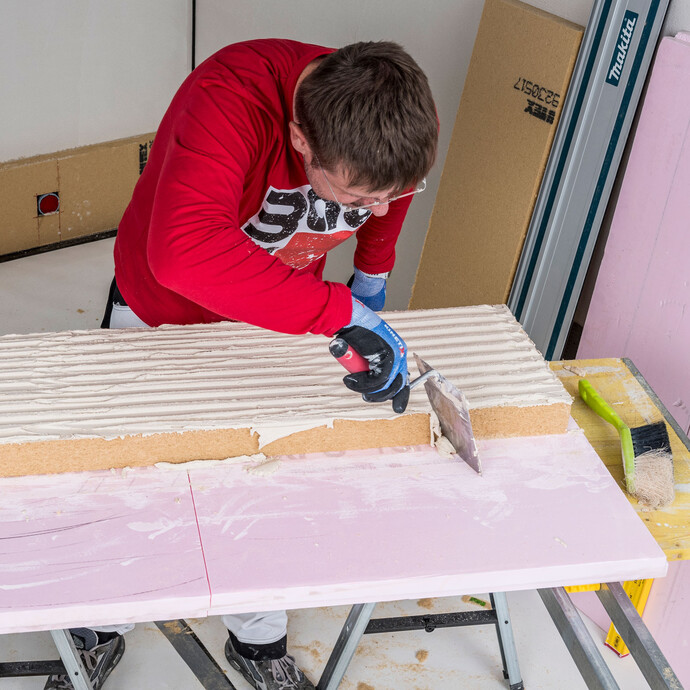 Heimwerker streift überstehenden Baumit Klebemörtel mit einer Maurerkelle umlaufend von der Holzfaserdämmplatte ab – saubere Plattenkanten vor der Verklebung der Innendämmung sicherstellen, Baumit Selbermachen Anleitung.